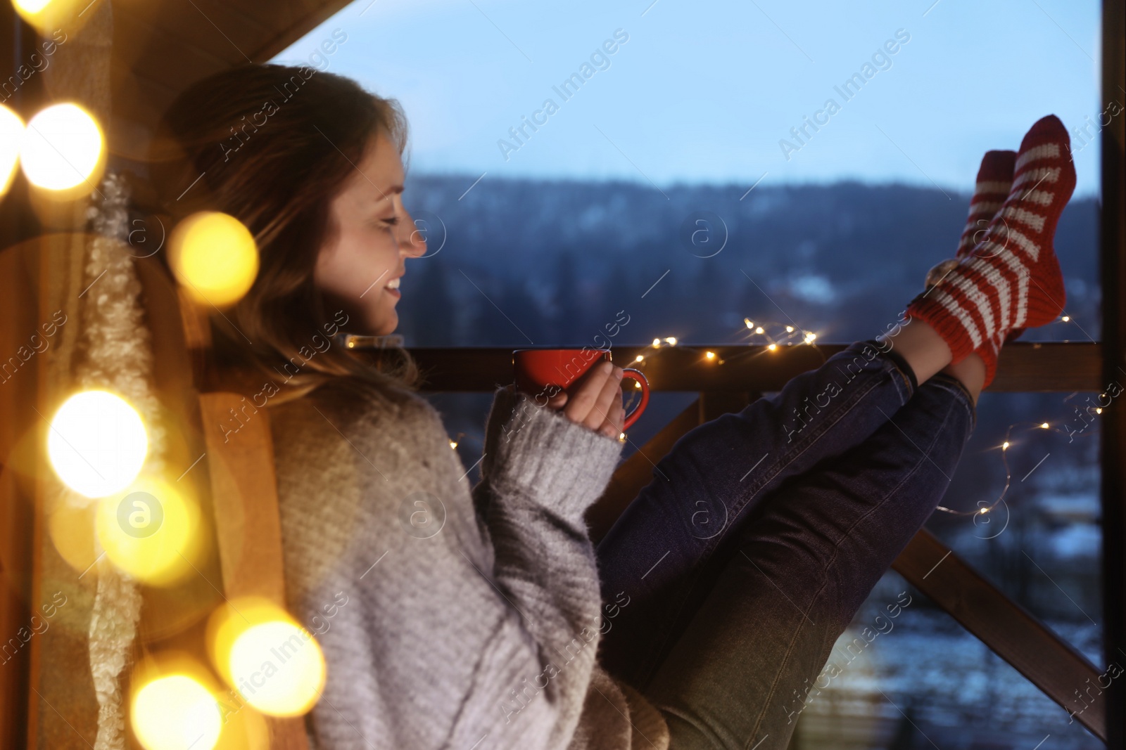 Photo of Woman with cup of hot beverage and Christmas lights resting on balcony. Winter evening