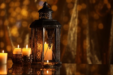 Photo of Arabic lantern and burning candles on mirror surface against blurred lights, space for text