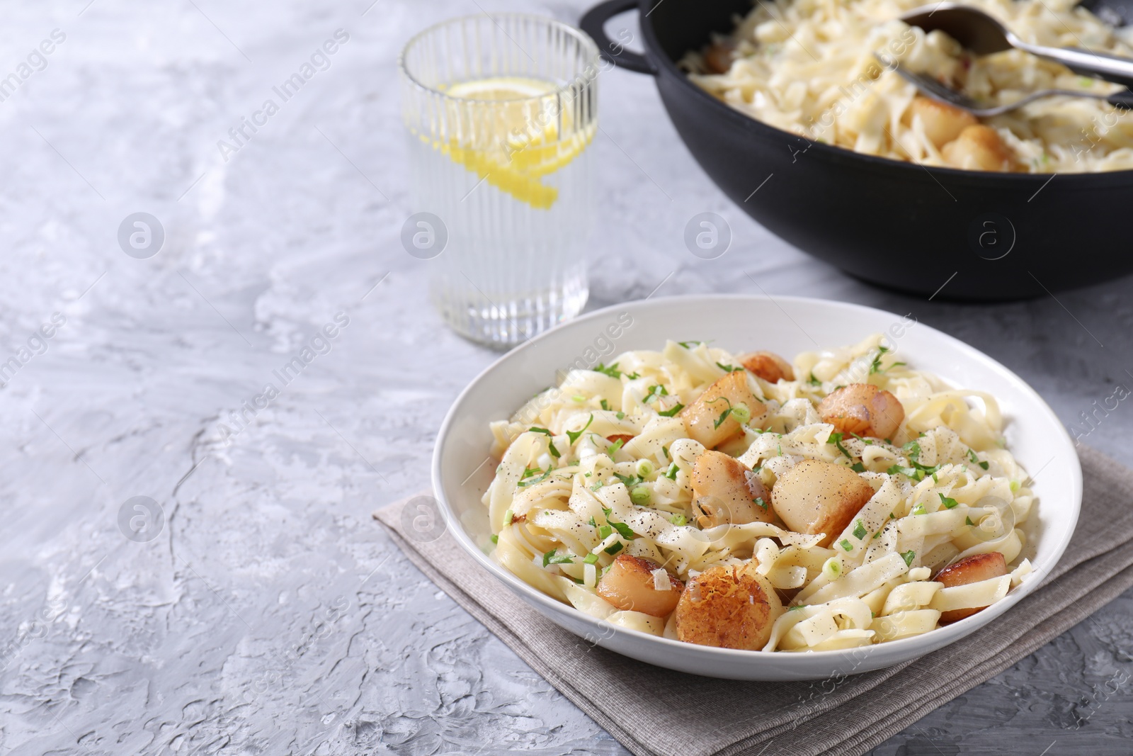 Photo of Delicious scallop pasta with spices in bowl and glass of water on gray textured table, closeup. Space for text