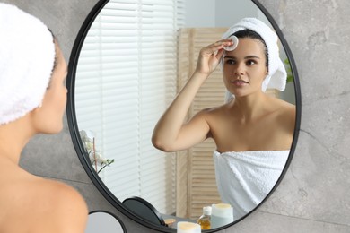 Photo of Young woman cleaning her face with cotton pad near mirror in bathroom