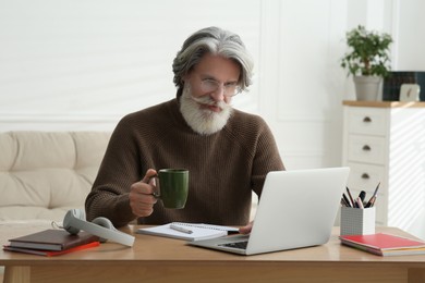 Photo of Middle aged man with laptop and cup of drink learning at table indoors