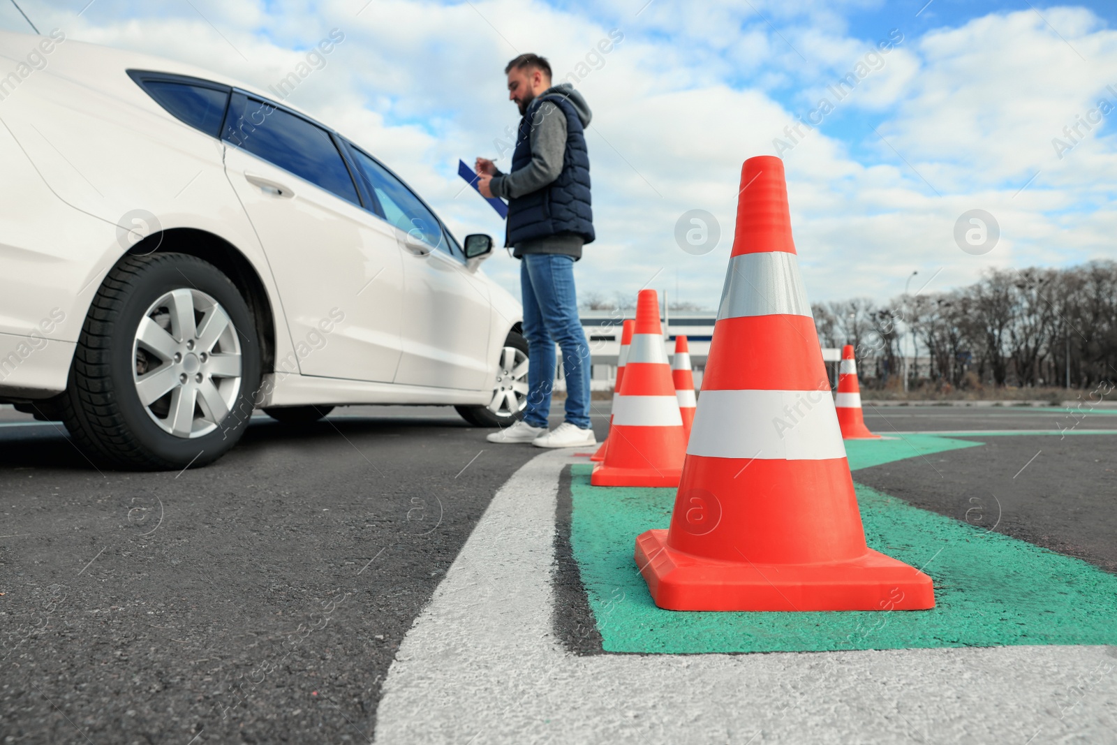 Photo of Instructor with clipboard near car outdoors, focus on traffic cone. Driving school exam