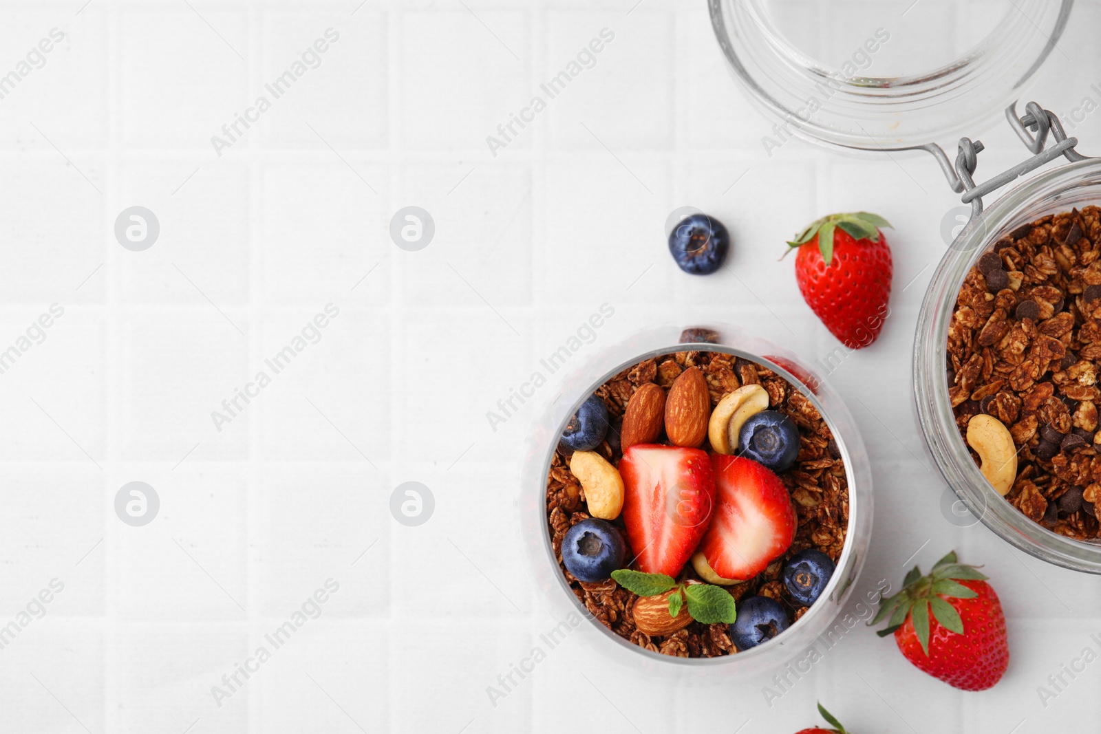 Photo of Tasty granola with berries, nuts and mint in glass on white tiled table, flat lay. Space for text