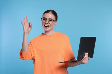 Happy woman with laptop showing okay gesture on light blue background