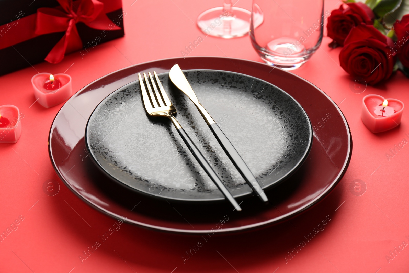 Photo of Place setting with heart shaped candles, gift box and bouquet of roses on red table, closeup. Romantic dinner