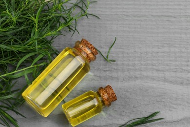 Bottles of essential oil and fresh tarragon leaves on grey wooden table, flat lay. Space for text