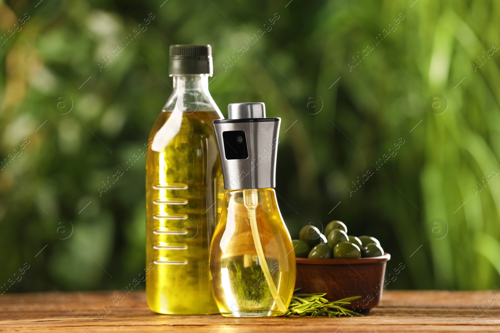 Photo of Bottles with cooking oil, olives and rosemary on wooden table against blurred background