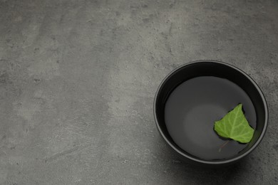 Photo of Black bowl with water and green leaf on grey table, above view. Space for text