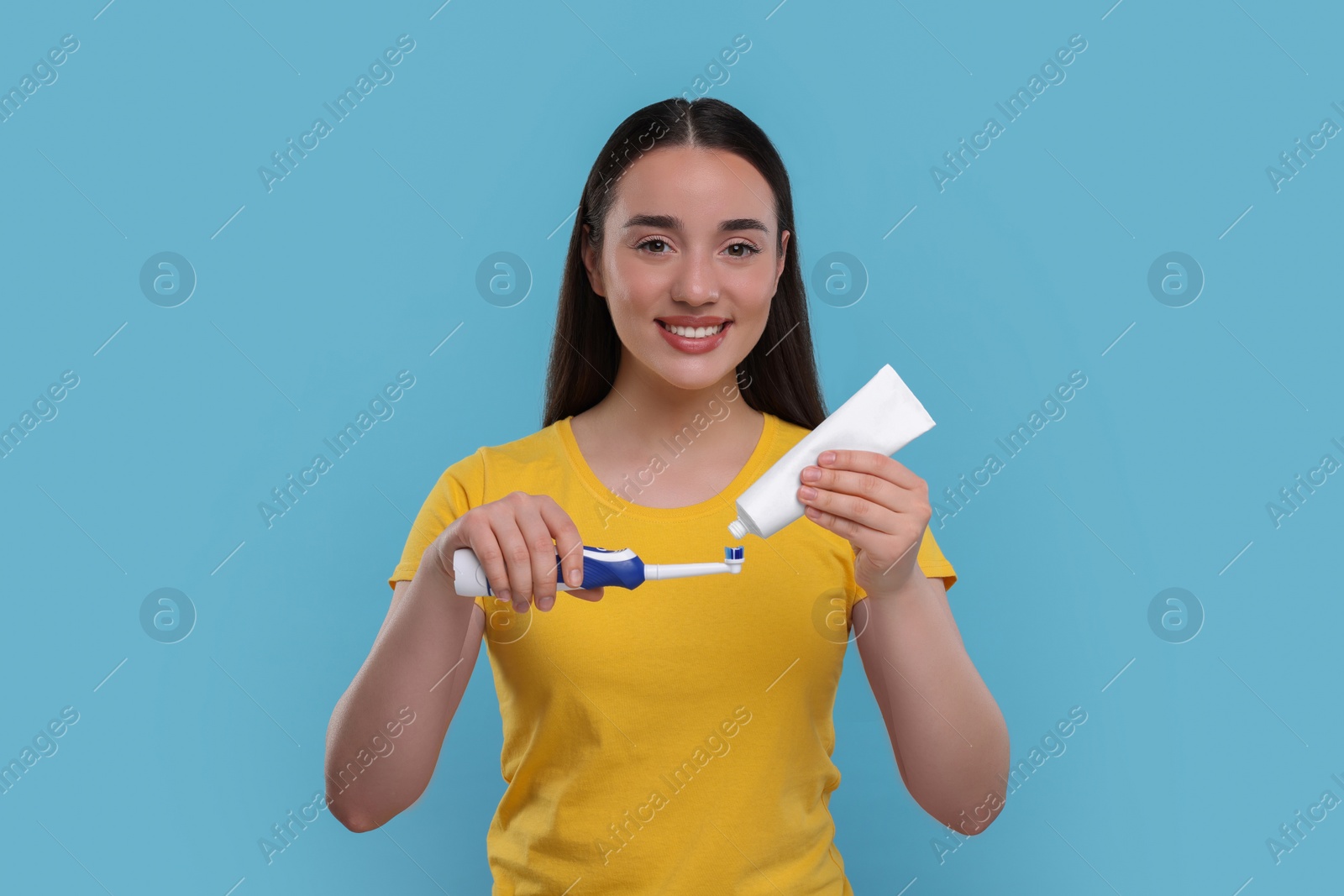 Photo of Happy young woman squeezing toothpaste from tube onto electric toothbrush on light blue background