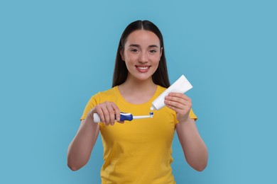 Happy young woman squeezing toothpaste from tube onto electric toothbrush on light blue background