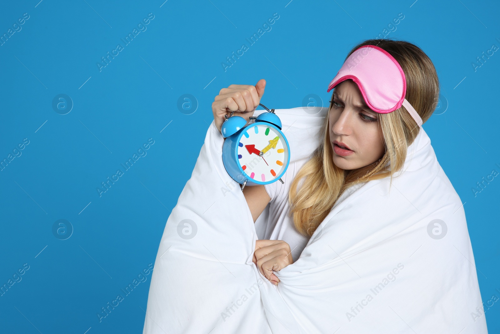 Photo of Emotional young woman in sleeping mask wrapped with blanket holding alarm clock on blue background