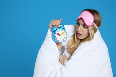 Photo of Emotional young woman in sleeping mask wrapped with blanket holding alarm clock on blue background