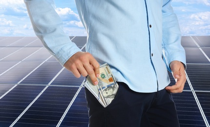 Image of Man with money and installed solar panels on background, closeup. Economic benefits of renewable energy