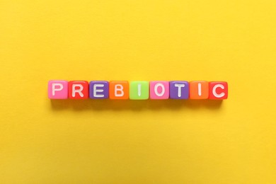 Photo of Colorful cubes with word Prebiotic on yellow background, flat lay