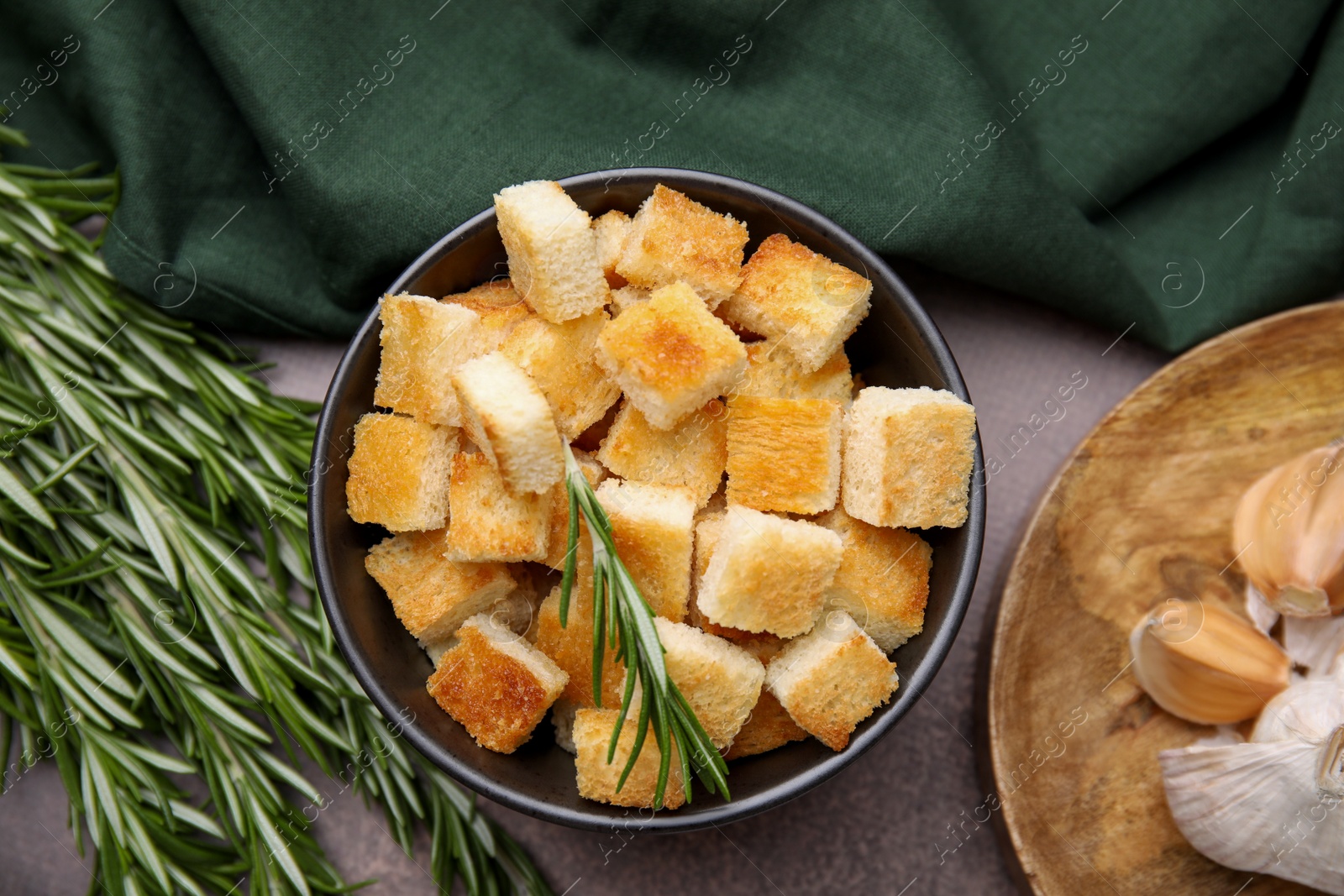 Photo of Delicious crispy croutons in bowl and rosemary on dark table, flat lay