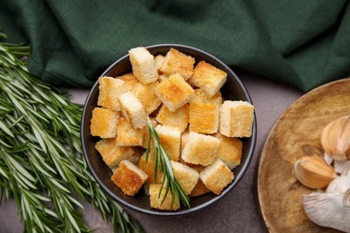 Photo of Delicious crispy croutons in bowl and rosemary on dark table, flat lay