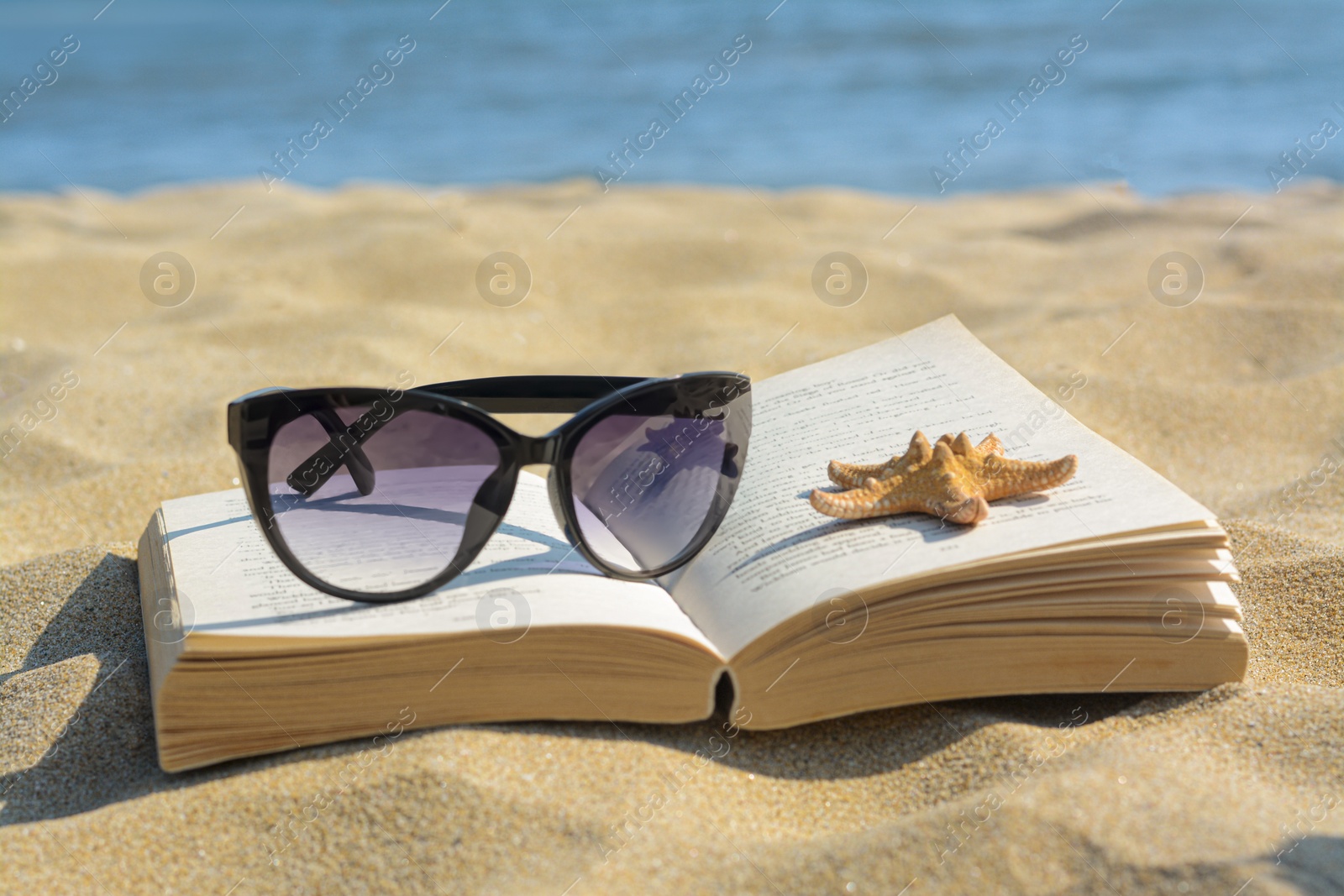 Photo of Beautiful sunglasses, book and starfish on sand near sea, closeup