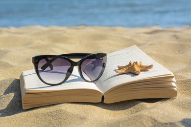 Photo of Beautiful sunglasses, book and starfish on sand near sea, closeup