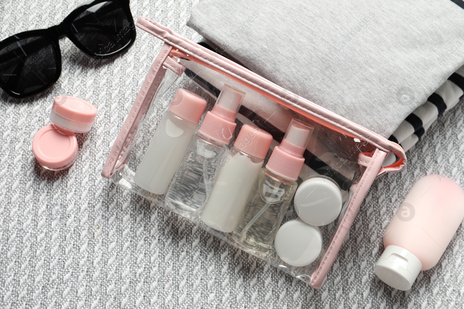 Photo of Cosmetic travel kit. Plastic bag with small containers of personal care products, stack of clothes and sunglasses on sofa