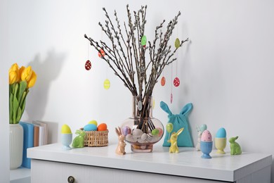 Beautiful pussy willow branches with paper eggs in vase and Easter decor on white chest of drawers at home