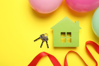 Wooden house model, keys, red ribbon and balloons on yellow background, flat lay with space for text. Housewarming party