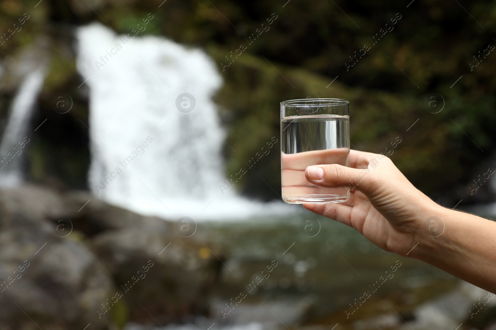 Photo of Woman holding glass of fresh water near waterfall outdoors, closeup. Space for text