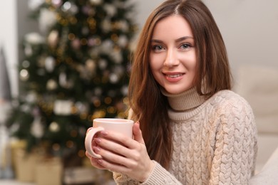 Photo of Beautiful young smiling woman with cup of tea near Christmas tree at home, space for text