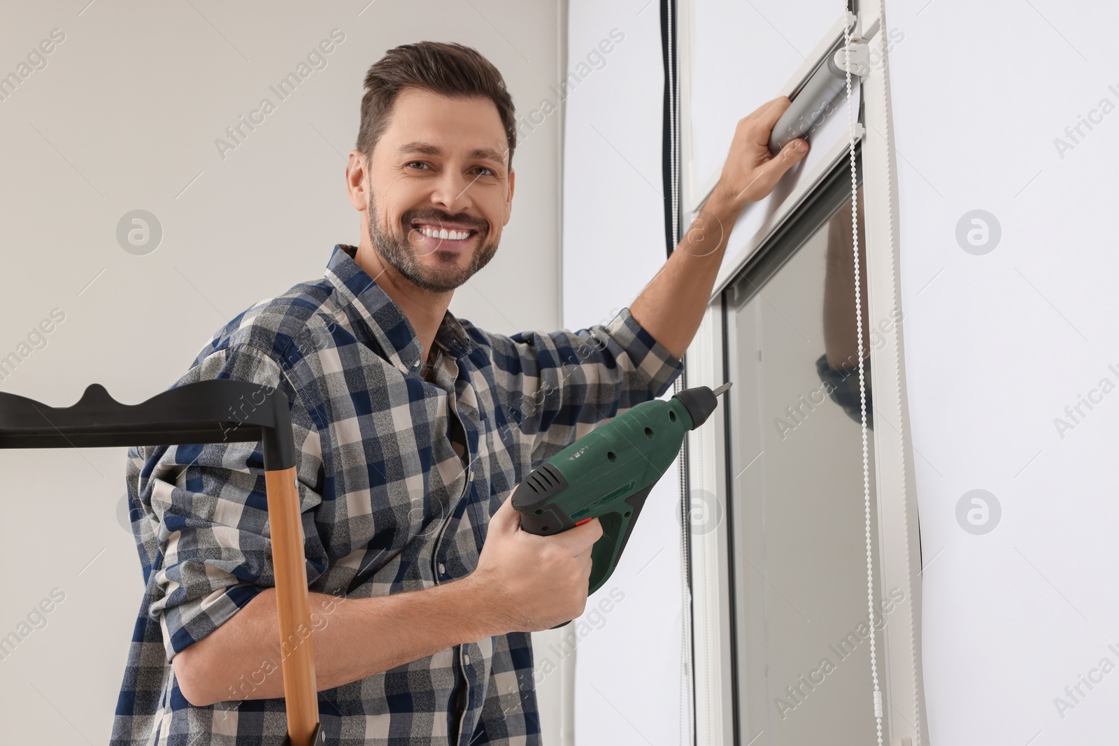 Photo of Man with drill installing roller window blind on stepladder indoors