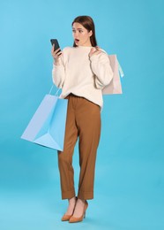 Photo of Surprised young woman with shopping bags looking at smartphone on light blue background. Big sale