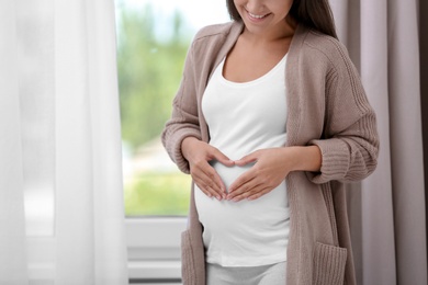 Pregnant woman standing near window at home, closeup