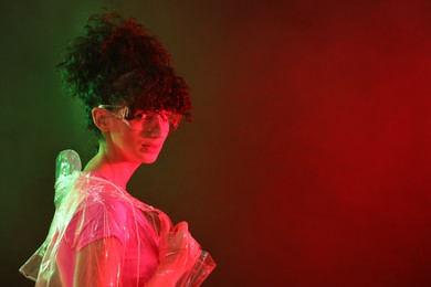 Photo of Beautiful young woman in transparent coat and sunglasses posing on color background in neon lights. Space for text