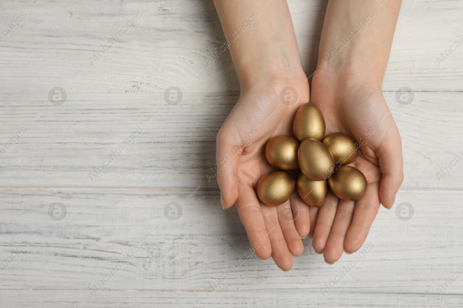 Photo of Woman holding shiny golden eggs at white wooden table, top view. Space for text