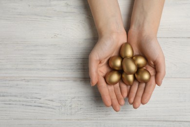 Woman holding shiny golden eggs at white wooden table, top view. Space for text