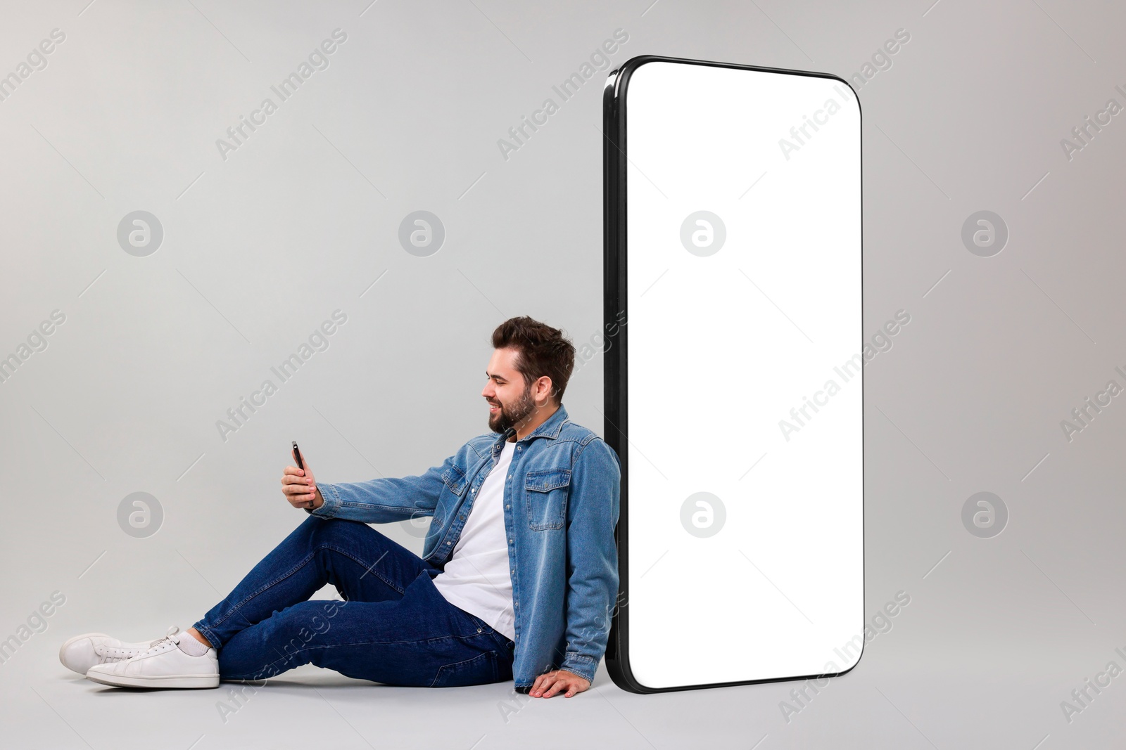 Image of Man with mobile phone sitting near huge device with empty screen on grey background. Mockup for design