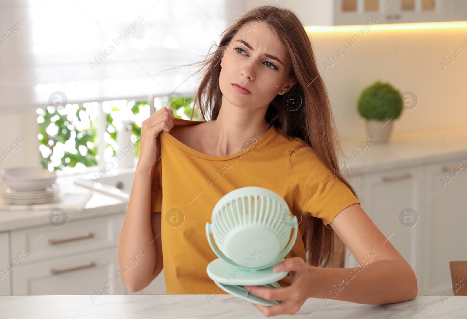 Photo of Woman with portable fan at table in kitchen. Summer heat