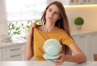 Woman with portable fan at table in kitchen. Summer heat