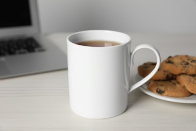 Photo of One ceramic mug with drink, laptop and tasty biscuits on white wooden table, closeup