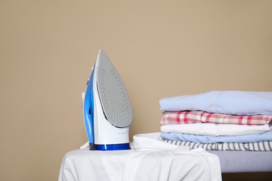 Photo of Modern iron, clean shirt and folded clothes on board against beige background