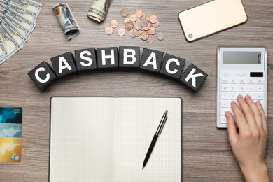 Photo of Woman using calculator at table with word Cashback made of black cubes, top view