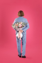 Photo of Man in stylish suit hiding beautiful flower bouquet behind his back on pink background
