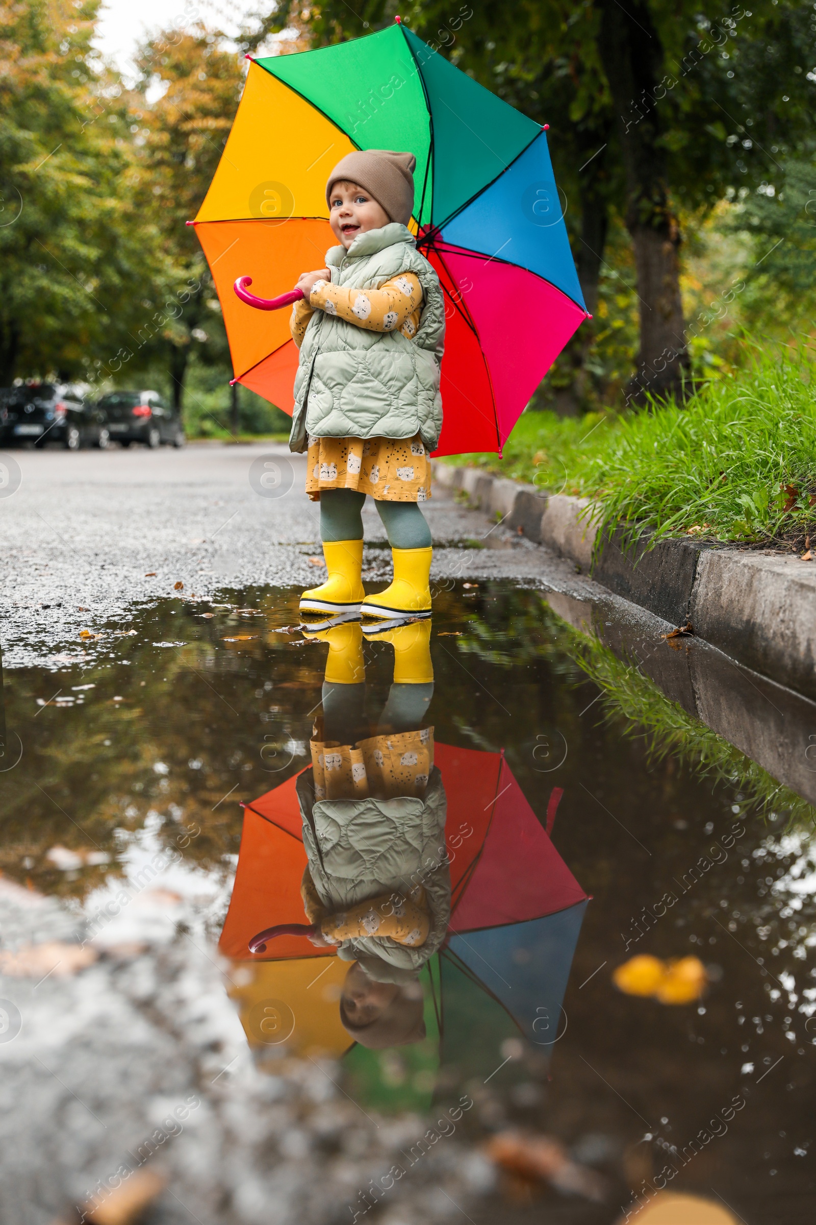 Photo of Cute little girl with colorful umbrella standing in puddle outdoors