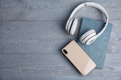 Book, modern headphones and smartphone on grey wooden table, flat lay. Space for text