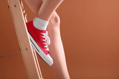 Photo of Woman in red classic old school sneakers on ladder against brown background, closeup. Space for text