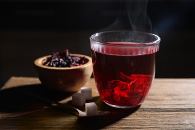 Photo of Delicious hibiscus tea in glass, sugar cubes and dry roselle petals on wooden table, closeup