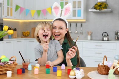 Photo of Mother and her son having fun while painting Easter eggs at table in kitchen