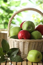 Photo of Different wet apples in wicker basket and green leaves on wooden table outdoors