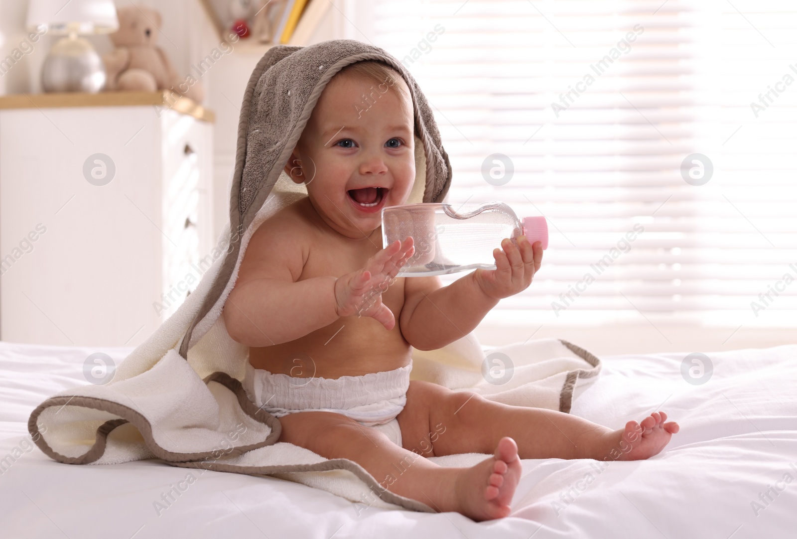 Photo of Cute little baby with towel and bottle of massage oil on bed at home