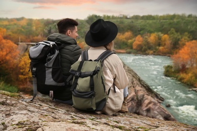 Photo of Couple of hikers with travel backpacks sitting on steep cliff near mountain river