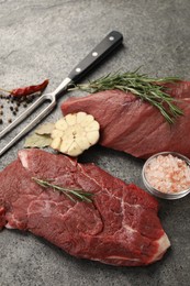 Piece of raw beef meat, spices, rosemary and fork on grey table