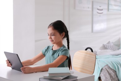 Photo of Little girl doing homework with tablet at table in room